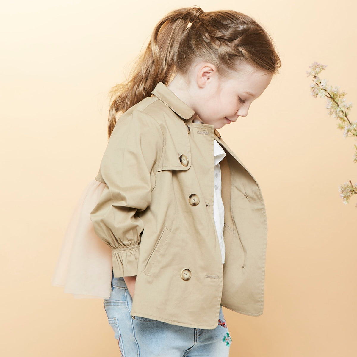 A trench coat, with a pink tutu sewn at the back. Two pockets are at the front and a buttoned panel on the upper left corner. Fastened from the front with faded dark/light brown coat buttons. 