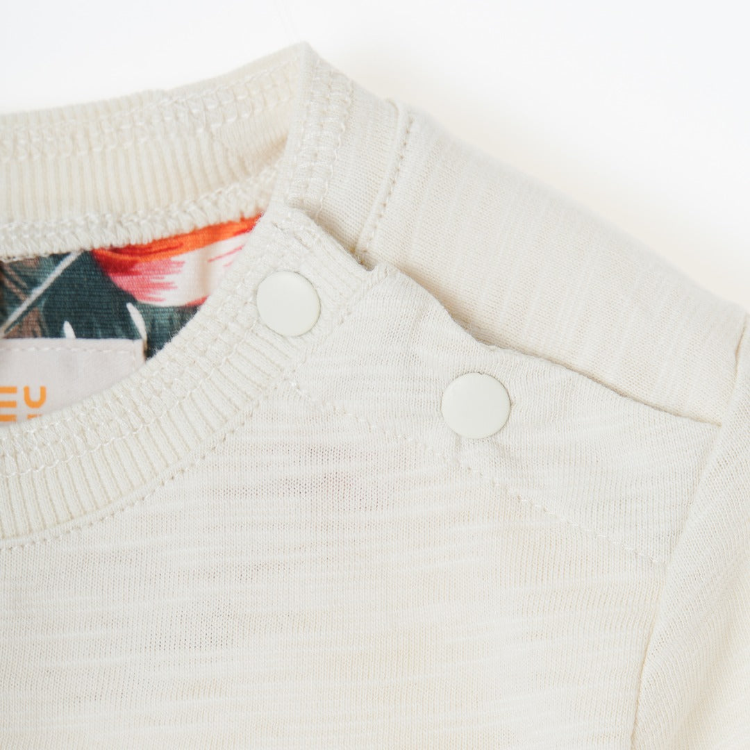 Cream colored, long sleeve top, hidden floral strap visible at the nape, front pocket and backside hem. 