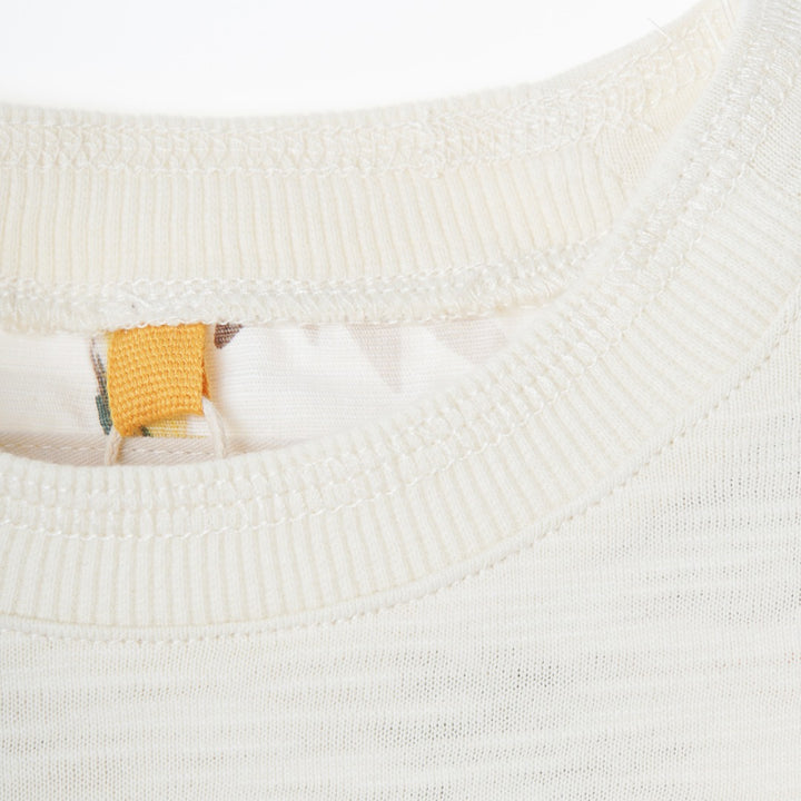 Cream colored, long sleeve top, hidden floral strap visible at the nape, front pocket and backside hem. 