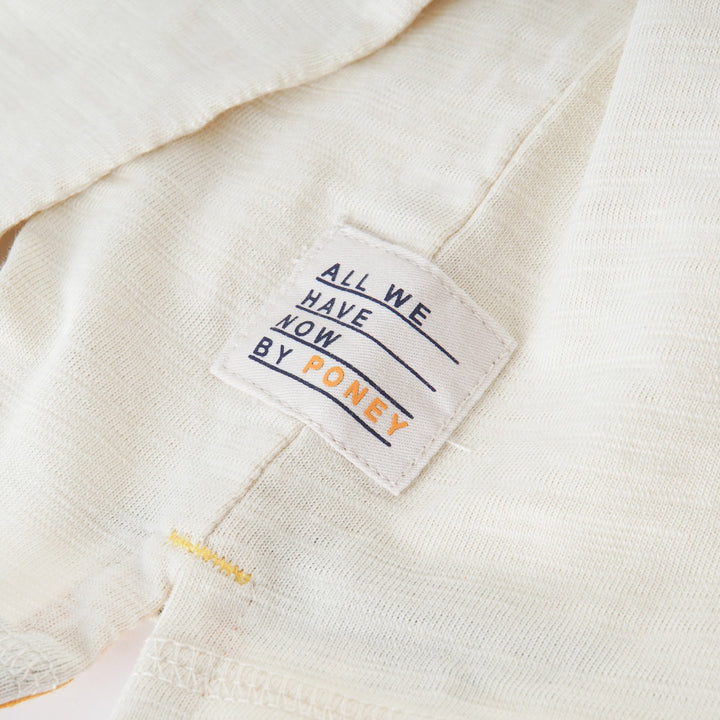 Cream colored, long sleeve top, hidden floral strap visible at the nape, front pocket and backside hem. 